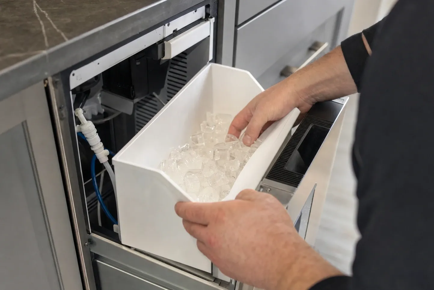 Hands lifting a bin of ice cubes out of an undercounter ice maker, with the inline water filter and supply tubing visible behind it