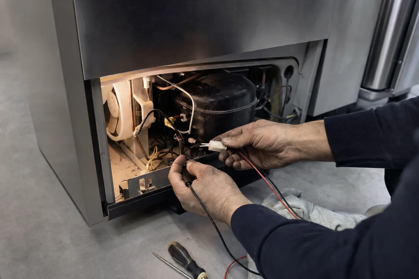 Multimeter probes held on a wiring connector beside a refrigerator compressor and condenser fan, the lower panel removed
