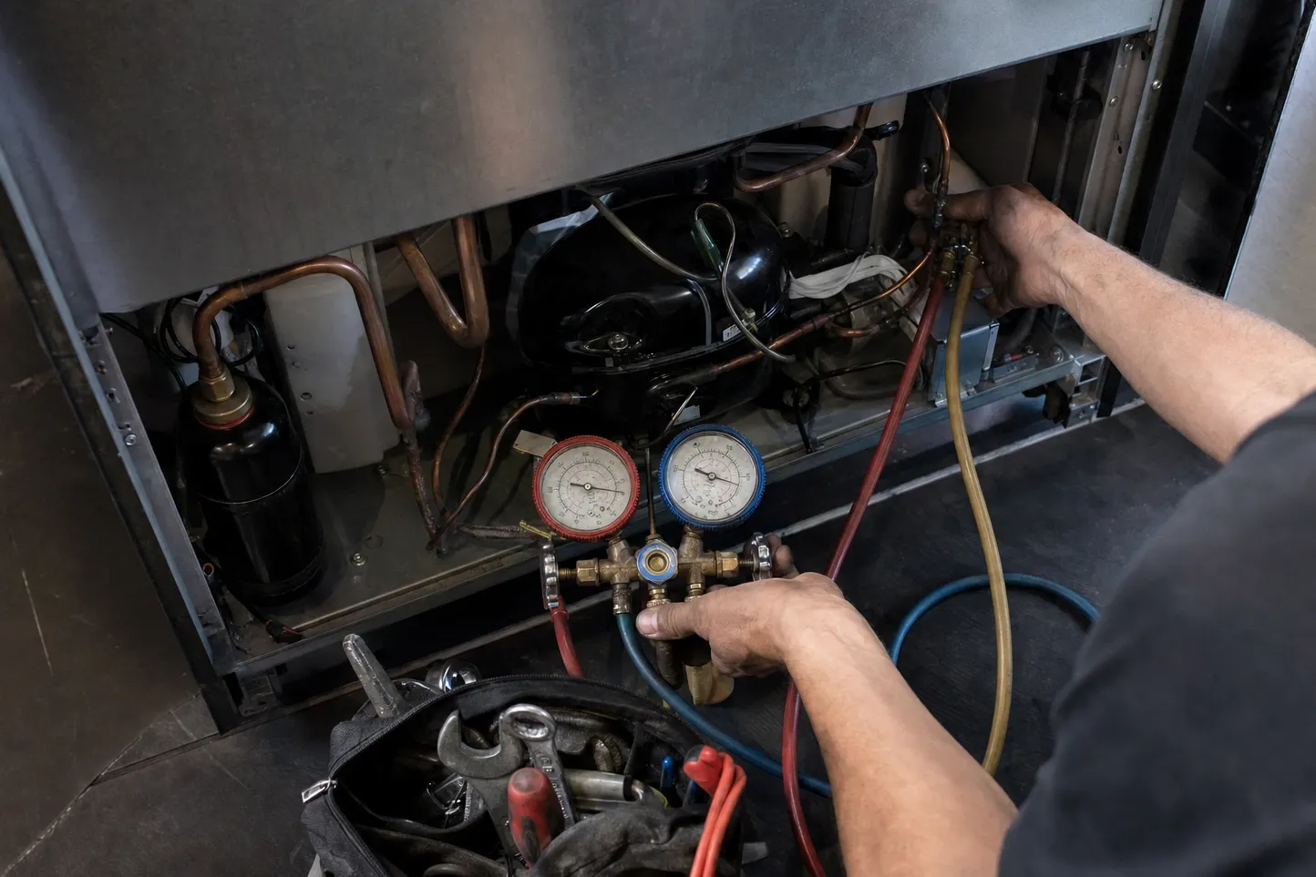 Manifold gauges connected at an open compressor bay seen from above, hands on the hose coupling and the valve knobs, copper lines around the compressor dome